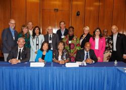 15 adults, all professionally dressed, gather for a group photo behind a conference table in a room with wood paneling. 