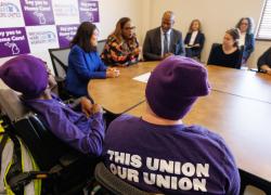 Acting Secretary Su sits around a table with a group of workers wearing purple and local leaders. The back of one worker’s shirt says, “This union, our union.” A banner behind her reads, “Say yes to Home care!” and “Michigan Home Care Workers United.”