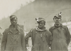 Three Black miners stand above ground, wearing coveralls and covered in coal dust.