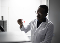 A black scientist in a white lab coat examines a test tube.