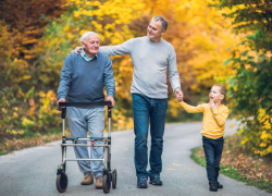 An elderly man, an adult man and a little girl walk down a road with trees with autumn foliage. The elderly man uses a walker.