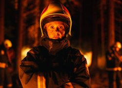 A female firefighter wearing her uniform and protective gear battling a fire in a smoky forest.