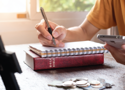 A person takes notes while using a calculator. A bible and cross are visible on the table.