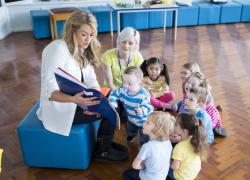 A woman shows a book to a group of toddlers while another woman looks on. They appear to be in a school or daycare setting.