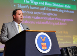 The image shows a person standing at a podium with the U.S. Department of Labor seal on it. The person is dressed in a suit and is delivering a speech. A presentation slide is visible in the background, related to human and labor trafficking topics.