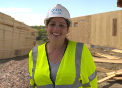 Clarissa Romero stands smiling at a construction site, wearing a hardhat labeled "OSHA" and a reflective vest.