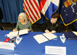 Dos mujeres están sentadas en una mesa firmando documentos. Las banderas de los Estados Unidos, la República Dominicana y el Departamento de Trabajo de los EE. UU. están detrás de ellas.