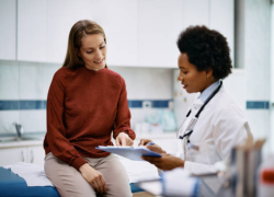 A white woman in a read turtleneck seated in a doctor's office with a Black woman doctor in a white coat and holding a clipboard.