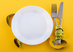 A white plate sitting next to silverware wrapped in yellow measuring tape placed on a yellow background.