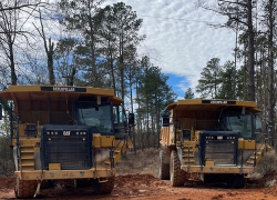 Two large dump trucks are parked in a wooded area with overhead power lines visible above them.