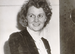 Sepia-toned photo of Elizabeth Hayes carrying a coat and purse. She has short, curly hair and is wearing a decorative jacket over a collared blouse.
