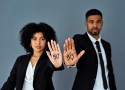 A man and woman in business suits stand side by side, holding out their hands. On both palms is written the number 50, but the 0 on the woman's hand is the male gender symbol, and the zero on the man's hand is the female symbol.