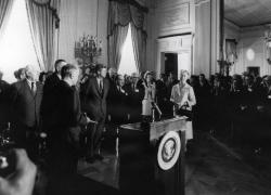 Black-and-white photo. Esther Peterson, President John F. Kennedy and dozens of others others stand in the White House. A podium is in the foreground.