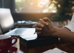 Close-up of a desktop with a laptop, mug and bible. A person sits with their hands clasped over the bible.
