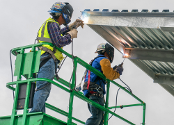 Two workers stand on an elevated platform to weld a metal roof. They are wearing fall protection harnesses, welding helmets, gloves and high visibility clothing.