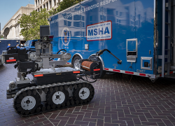 A robot with tracks like a bulldozer and a variety of mechanical parts sits outside a large blue trailer with MSHA's logo and "Mine Emergency Operations Robotic Response Team" 