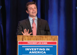 A man in a suit speaks at a podium labeled "President Joe Biden - Investing in America"