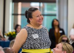 Una mujer sonriente con un identificador de nombre en el que se lee “Kimberly Rush” se encuentra de pie en un cuarto de conferencias rodeada de otras mujeres. 