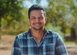 A man wearing a blue checkered button-down collared shirt smiles at the camera while standing in an open field with trees behind him.