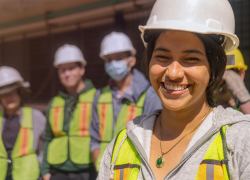 Uma jovem trabalhadora usando um capacete de proteção sorri, com vários outros trabalhadores em pé atrás dela.