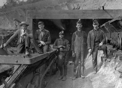 Five miners stand or sit on equipment at the entrance of a coal mine. The man in the middle is Black and the other four are white. Library of Congress photo.