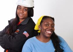 A Black woman with long hair standing while wearing a black jacket and white hardhat and a Black woman with braids wearing a yellow scarf and blue t-shirt resting her arm on a white hardhat.