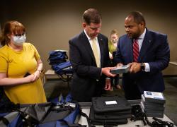 Dallas Mayor Eric Johnson passes a Mi-Fi to Secretary Marty Walsh to place in a package at the J. Erik Jonsson Central Library. To the Secretary's left is an employee of the library. In front of the Secretary is a table of digital devices available for check out.  
