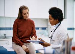 A woman wearing a rust-colored turtleneck and khaki pants reviews medical documents on a clipboard with a doctor wearing a white coat and stethoscope. 