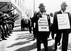 Black sanitation workers participate in a strike in Memphis in 1968 as National Guard members look on. One worker carries a sign that reads “End dismal working conditions now.” 