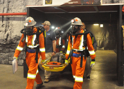 A mine rescue team wearing orange safety suits and respirators emerges from a tunnel carrying a stretcher after completing an exercise as part of the International Mine Rescue Competition.
