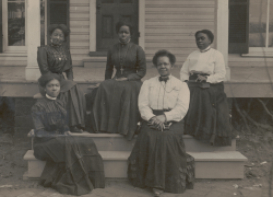 Nannie Helen Burroughs sits with four other Black women in Victorian dress on the steps to the front porch of a building.