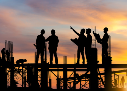 Silhouettes of construction workers standing on scaffolding as the sun sets behind them.