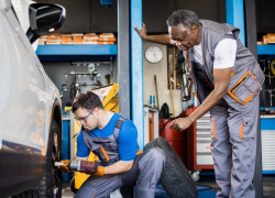 In a garage, a young man with Downs syndrome works on a car tire under the supervision of an adult mentor.