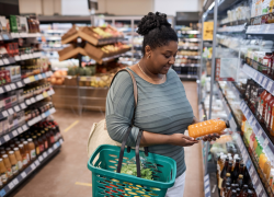 A woman holding a shopping basket looks at a bottle of juice in a grocery store refrigerator aisle.