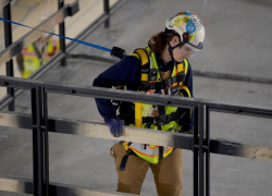 Isang babaeng manggagawa sa konstruksiyon na nakasuot ng mataas na visibility gear, isang helmet na pangkaligtasan na may chinstrap at malinaw na visor, isang harness na proteksyon sa pagkahulog at mga guwantes.