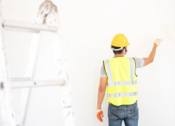 A worker wearing a high visibility vest and hardhat, seen from behind, touches up a white wall with a paint brush.