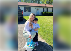 A woman crouches in a driveway to embrace a young boy. She holds an Easter basket full of colorful eggs.