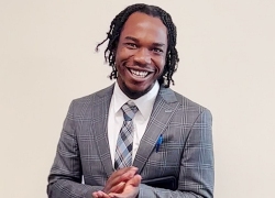 Photo of Tajay Kelly standing in a grey suit and smiling