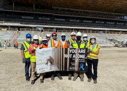 A group of Alberici Corp. workers participate in a 2021 Suicide Prevention Stand-Down at a Major League Soccer stadium construction site in St. Louis. They are holding a banner that says "You are not alone."