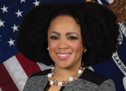 Pictured is a black woman with shoulder length natural hair smiling at the camera in front of the American and U.S. Department of Labor flags. 