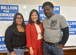 A woman with long dark hair wearing a black shirt and jeans stands with a woman with collar-length black hair wearing a red suit and a man with short dark hair wearing a grey t-shirt and jeans. They are smiling for a photo and standing in front of a white board with signs that read, " $1 BILLION FOR WORKERS RECOVERED."