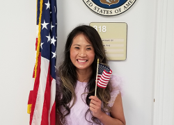 A woman in a pink dress poses by an American flag, holding a miniature flag and beaming at the camera. The Labor Department seal is visible behind her.