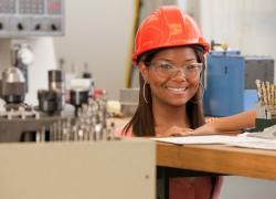 A young woman in a hardhat and safety goggles smiles while standing in a workshop.