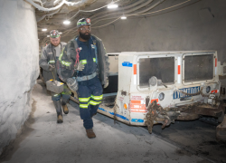 Two underground coal miners wearing hard hats with headlamps exit a transport vehicle and walk toward their work site.