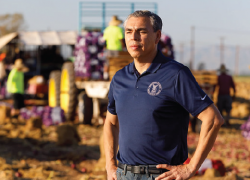 Wage and Hour investigator Juan Coria stands in a field. Workers are visible on a tractor behind him.