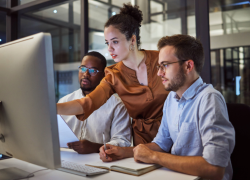 Three workers lean over a desk, looking at a computer screen.