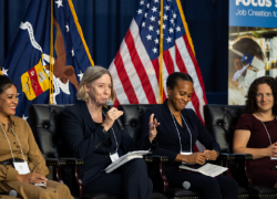 A seated woman in a business jacket addresses the audience. Three other women on the panel listen attentively.