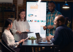 Several employees work around a table with laptops and tablets. A large screen with a data presentation is behind them.