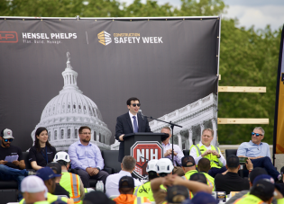 Deputy Secretary Keith Sonderling speaks at a construction safety event. A large backdrop behind him says "Hensel Phelps" and "Construction Safety Week." The podium has the National Institutes of Health logo.