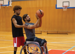 A personal trainer guides a man in a wheelchair as he prepares to shoot a basketball. Photo by Kampus Production.